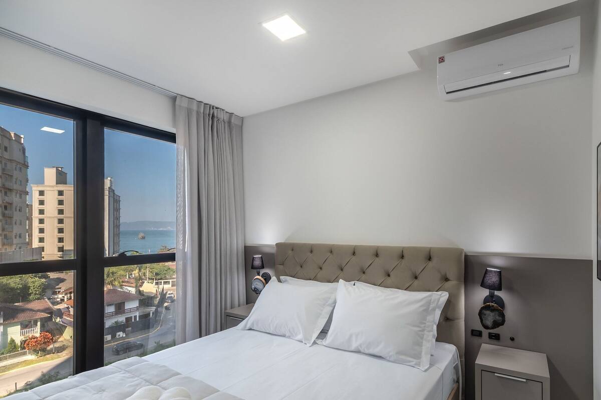 A hotel room with a large bed, a view of buildings and the sea, an air conditioning unit, and a nightstand with a lamp.