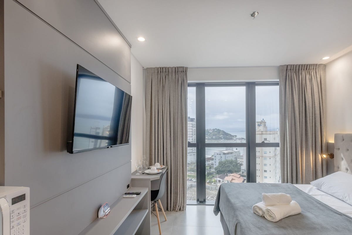 Ett modernt hotellrum med en säng, ett skrivbord med en stol, en tv som är monterad på väggen och ett stort fönster med utsikt över staden.