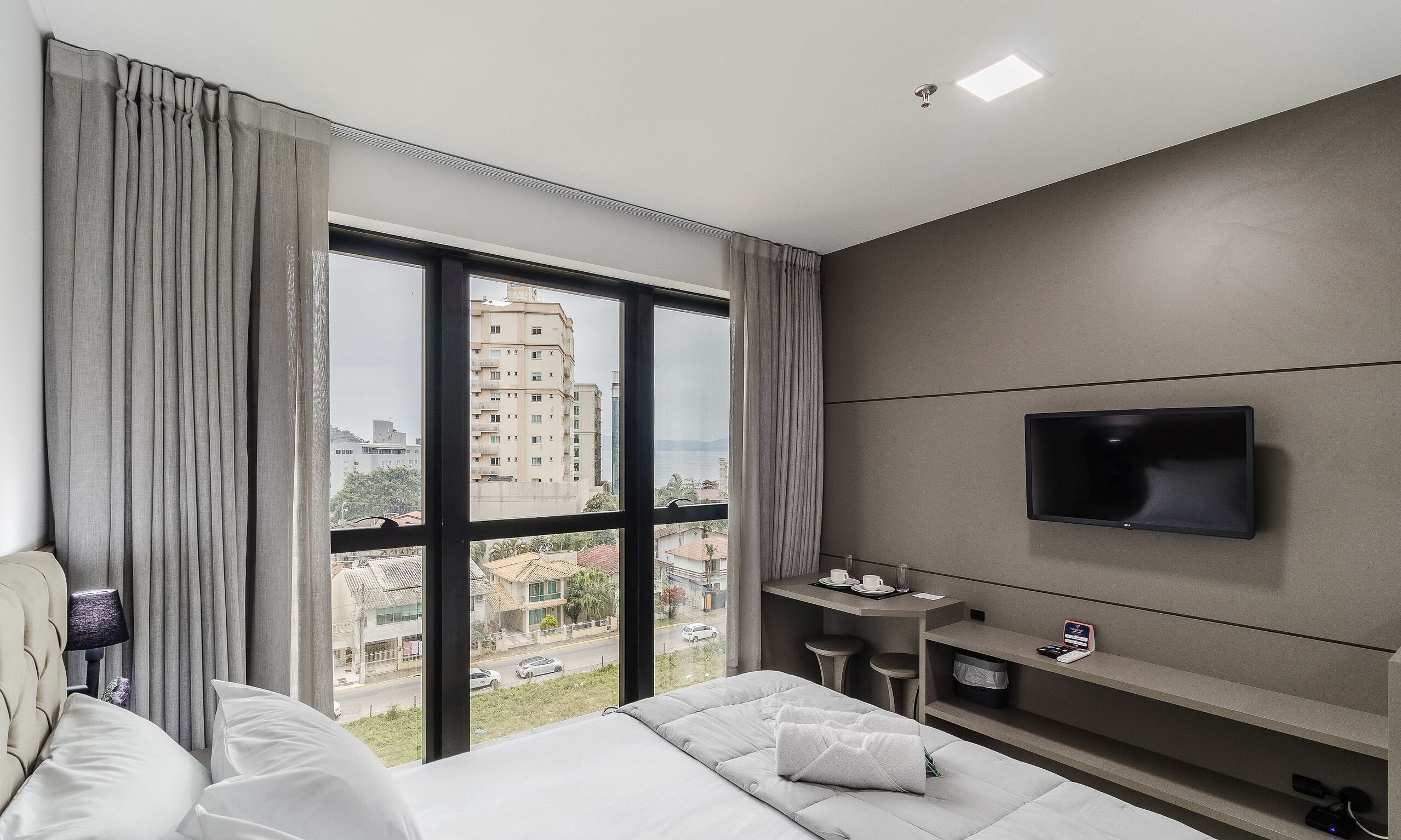 Une chambre d’hôtel avec un lit, un téléviseur fixé au mur, une petite table avec une tasse et une soucoupe, et une vue sur les bâtiments à travers de grandes fenêtres.