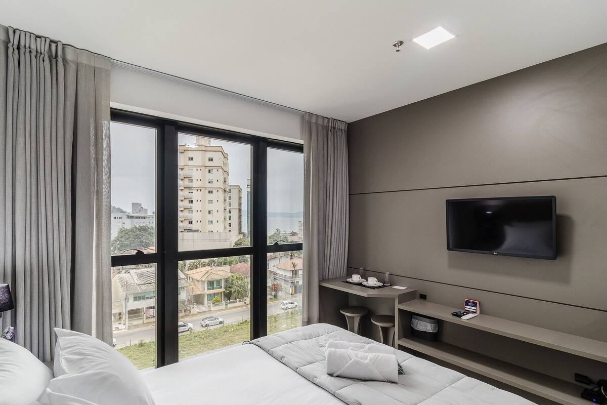 Une chambre d’hôtel avec un lit, un téléviseur fixé au mur, une petite table avec une tasse et une soucoupe, et une vue sur les bâtiments à travers de grandes fenêtres.