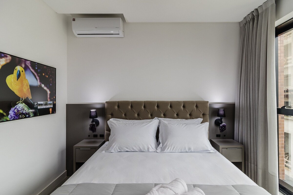 Habitación de hotel con cama, dos mesitas de noche, aire acondicionado instalado en la pared, ventana con cortinas e imagen enmarcada en la pared.