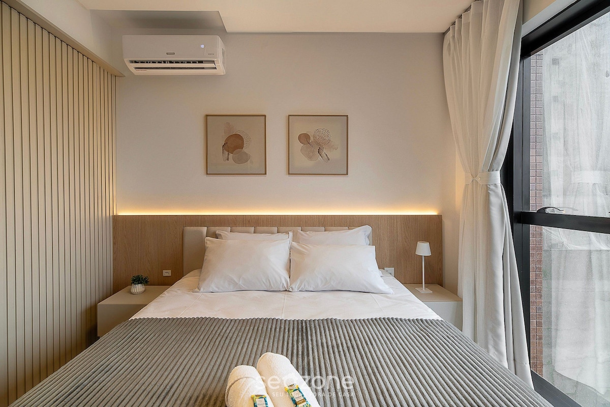 A hotel room with a bed, a nightstand, an air conditioner, and a window with curtains.