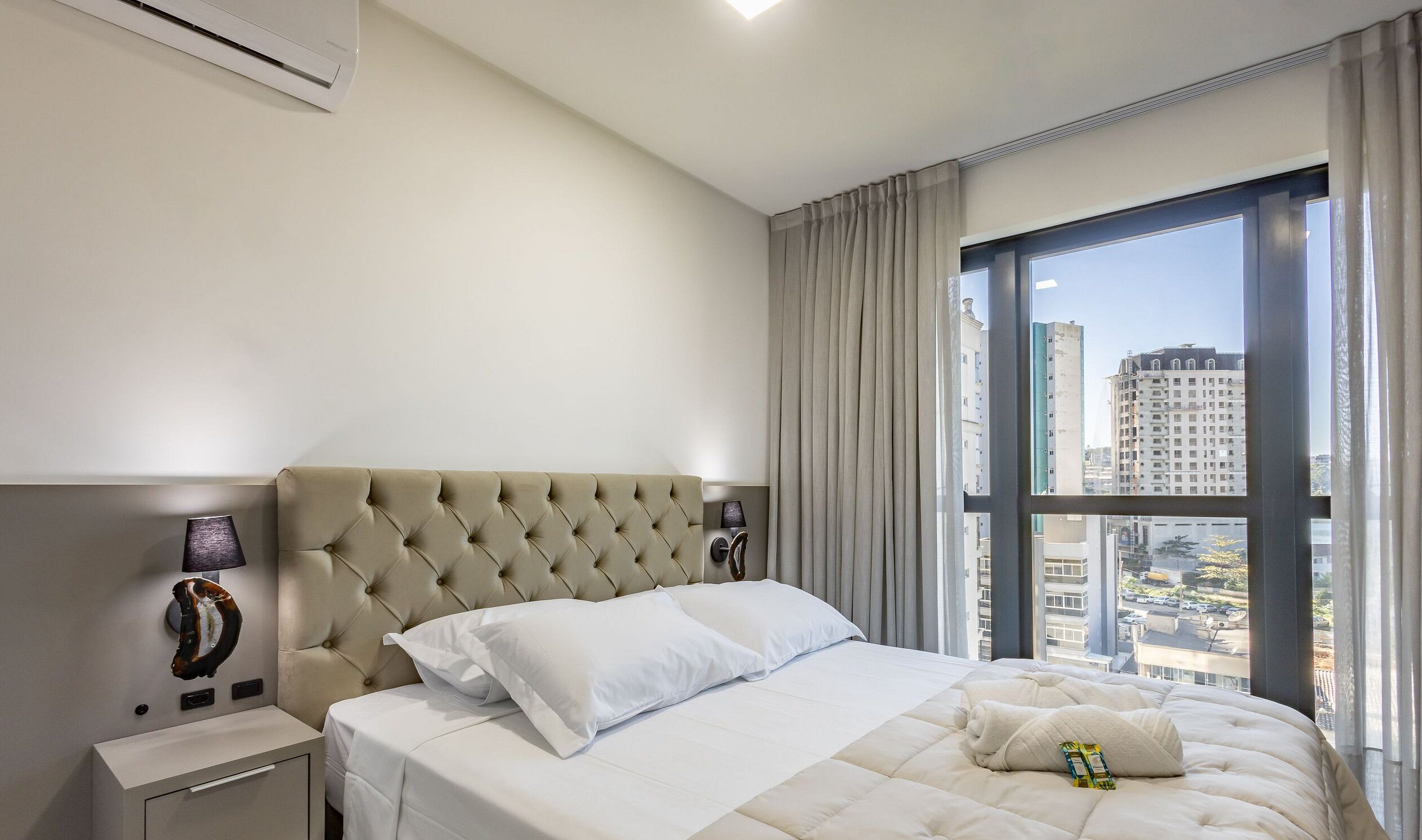 Une chambre d’hôtel avec un grand lit, une table de chevet, une lampe, une fenêtre avec des rideaux et une vue sur les bâtiments.
