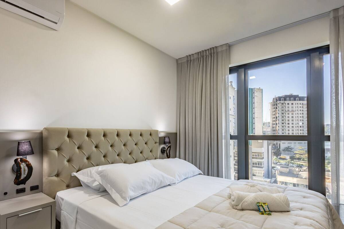 Une chambre d’hôtel avec un grand lit, une table de chevet, une lampe, une fenêtre avec des rideaux et une vue sur les bâtiments.