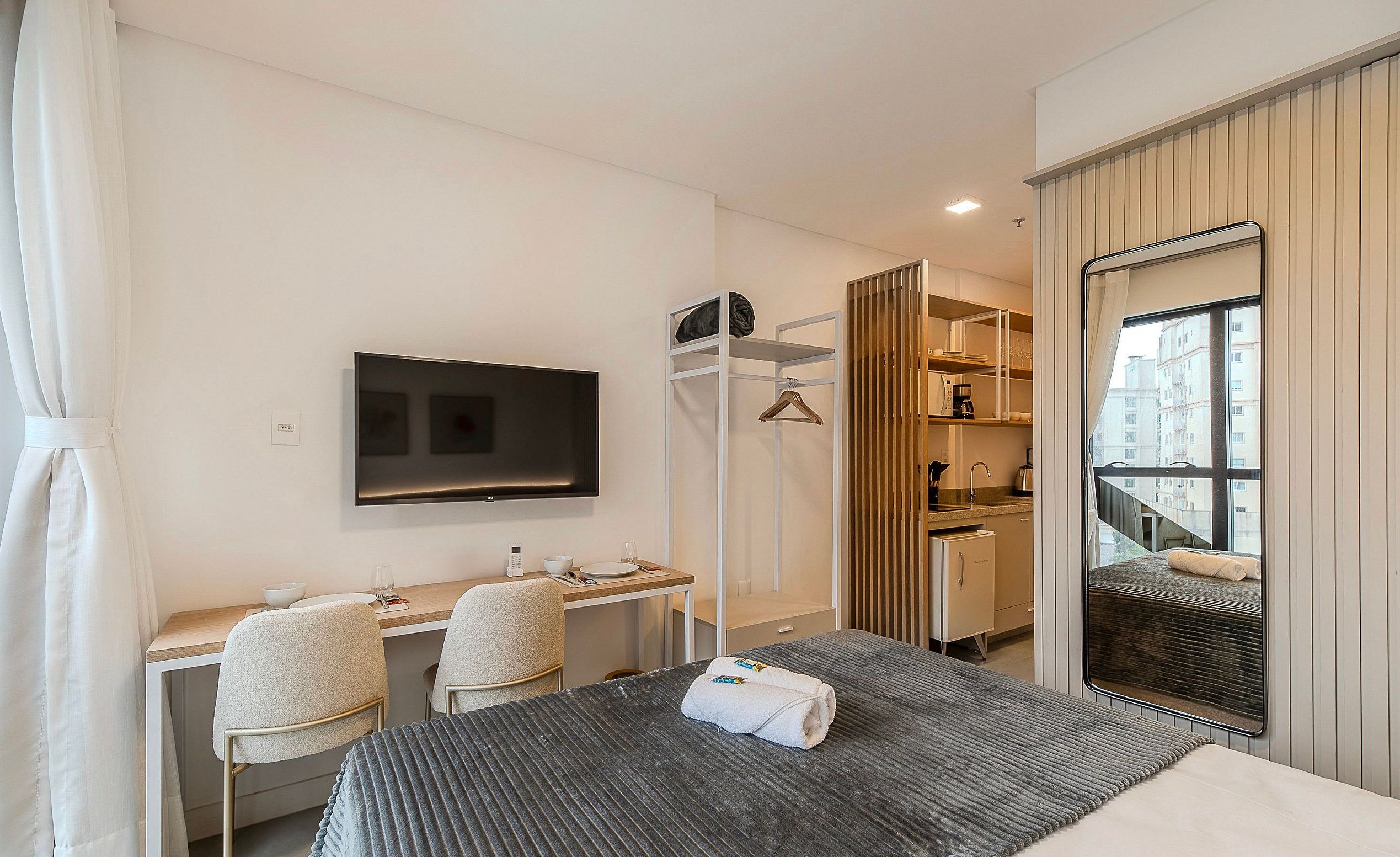 Habitación de hotel con cama, escritorio, dos sillas, televisor, armario y ventana con vista a edificios.