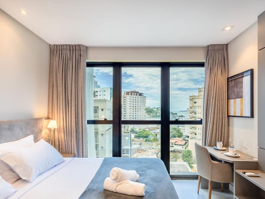 A hotel room with a bed, a desk, a chair, a TV, and a view of the cityscape.