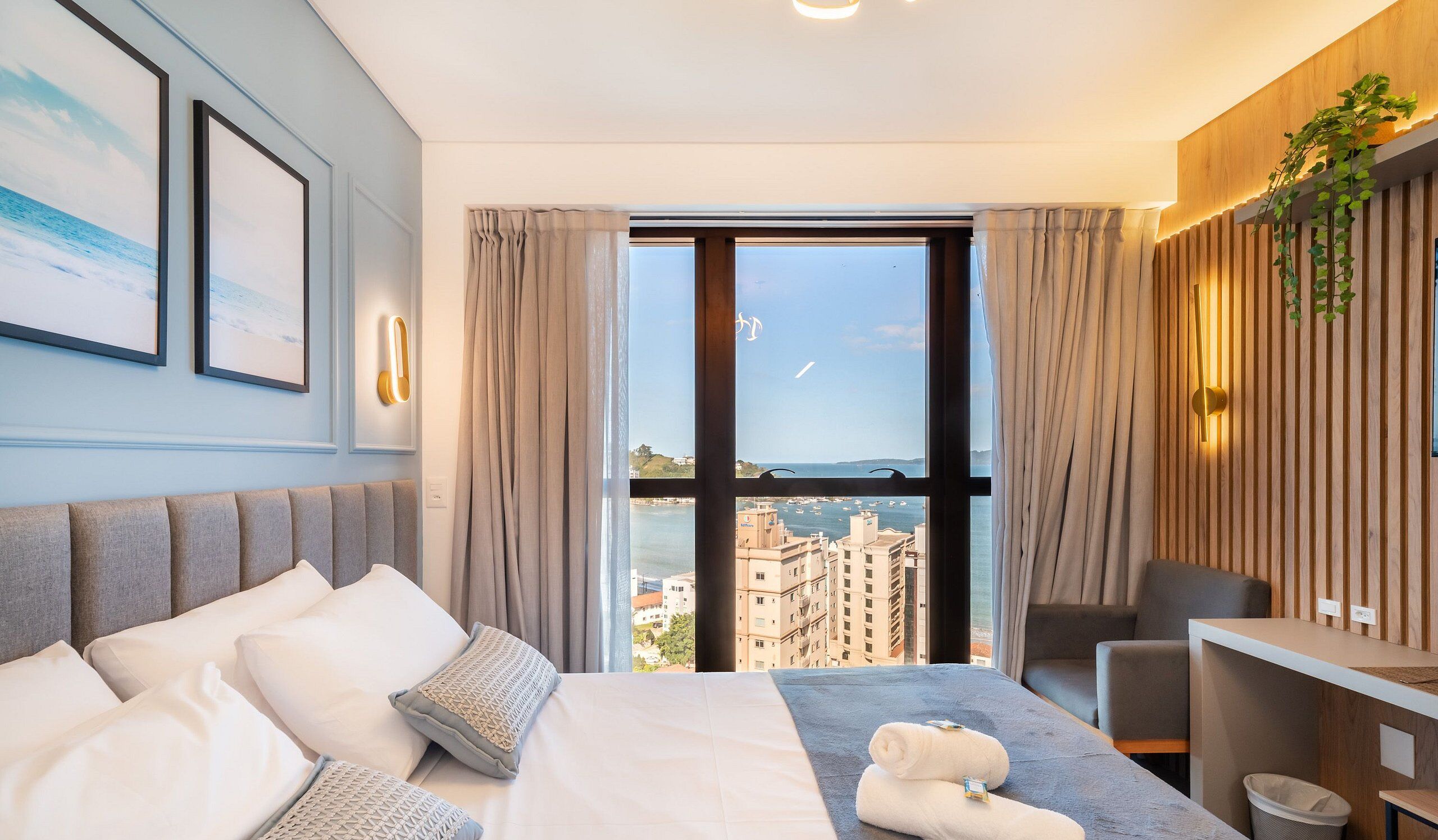 Une chambre d’hôtel comprenant un lit, un bureau, une chaise, une grande fenêtre donnant sur des bâtiments et sur l’eau, et une plante fixée au mur.