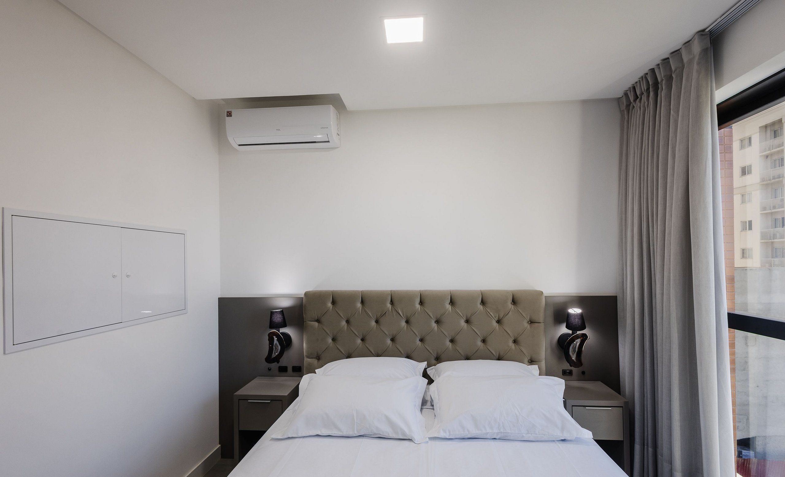 Una habitación de hotel con cama, dos lámparas de noche, aire acondicionado instalado en la pared y una ventana con cortinas.