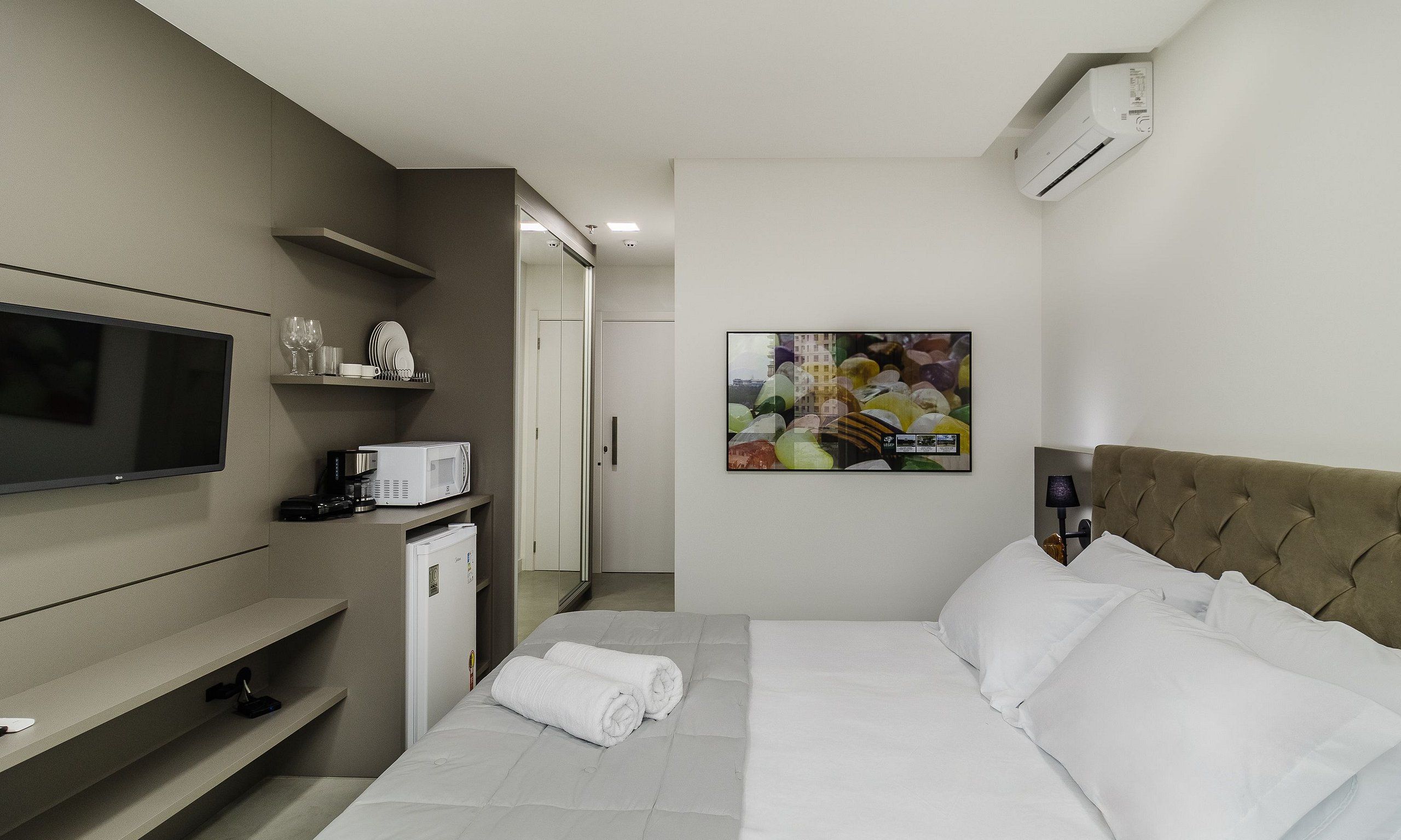 Una habitación de hotel con cama, televisor, microondas, cafetera, refrigerador pequeño, mesita de noche y aire acondicionado instalado en la pared.