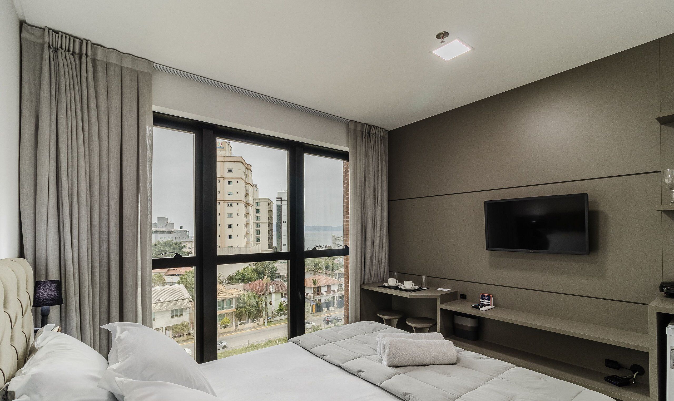 Une chambre d’hôtel moderne dotée d’un grand lit, d’un téléviseur fixé au mur, d’un bureau et offrant une vue sur le paysage urbain grâce à de grandes fenêtres.
