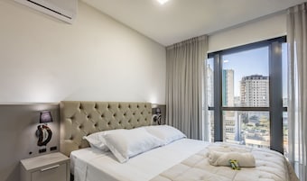 Habitación de hotel con una cama grande, una mesita de noche, una lámpara y vistas a la ciudad a través de un ventanal amplio.
