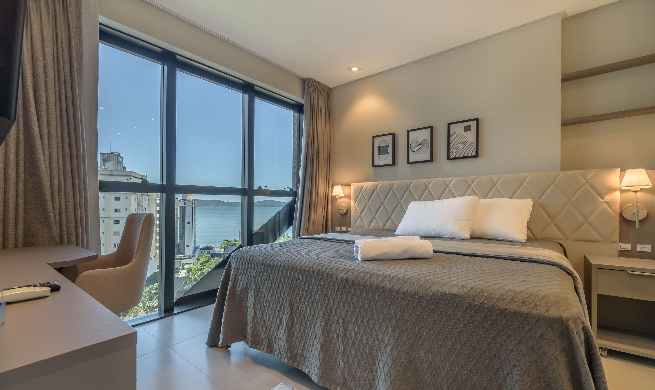 Une chambre d’hôtel dotée d’une grande fenêtre, d’un lit avec une tête de lit matelassée, d’un bureau avec une chaise et d’une vue sur la ville.