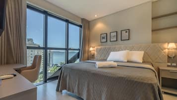 Quarto de hotel com uma cama grande, escrivaninha, cadeira, vista para o mar e obras de arte na parede.