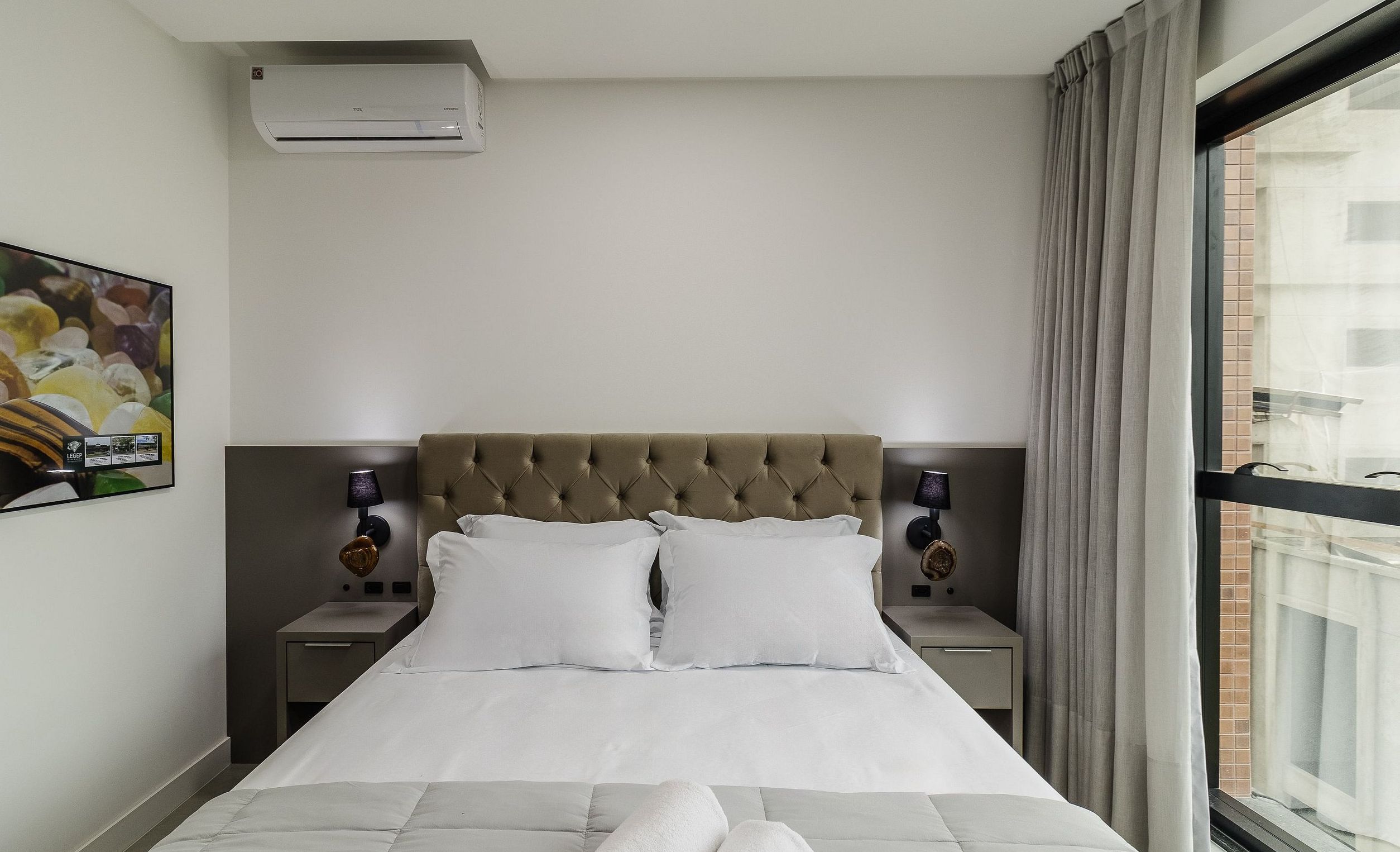 A hotel room with a bed, two bedside lamps, a wall-mounted air conditioner, and a window with curtains.