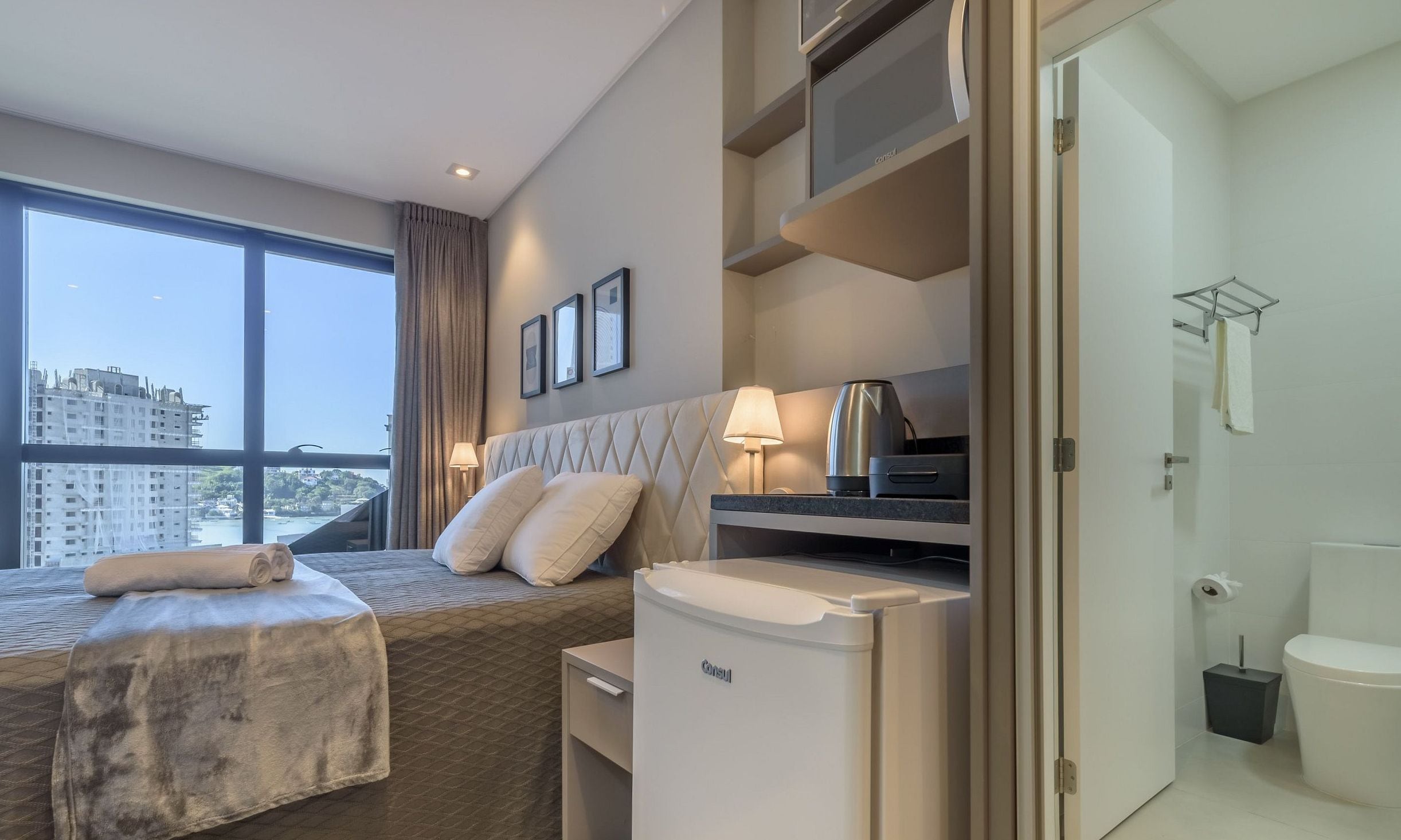 Ein Hotelzimmer mit Bett, Schreibtisch, Lampe, Minikühlschrank und Blick auf die Stadt.