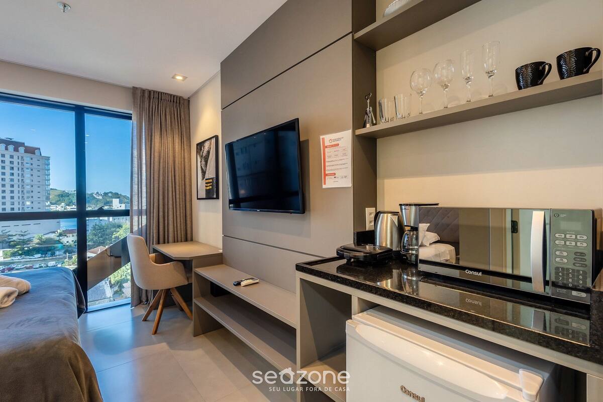 Une chambre d’hôtel moderne équipée d’un lit, d’un bureau, d’une télévision à écran plat, d’un minibar et d’une grande fenêtre donnant sur la ville.
