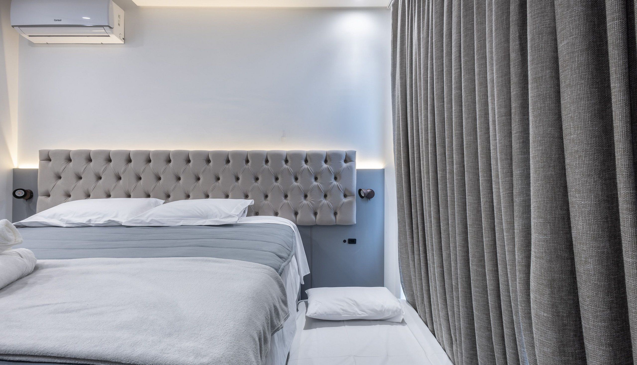 A hotel room with a bed, bedside tables, a nightstand, a wall-mounted air conditioner, and a window with curtains.