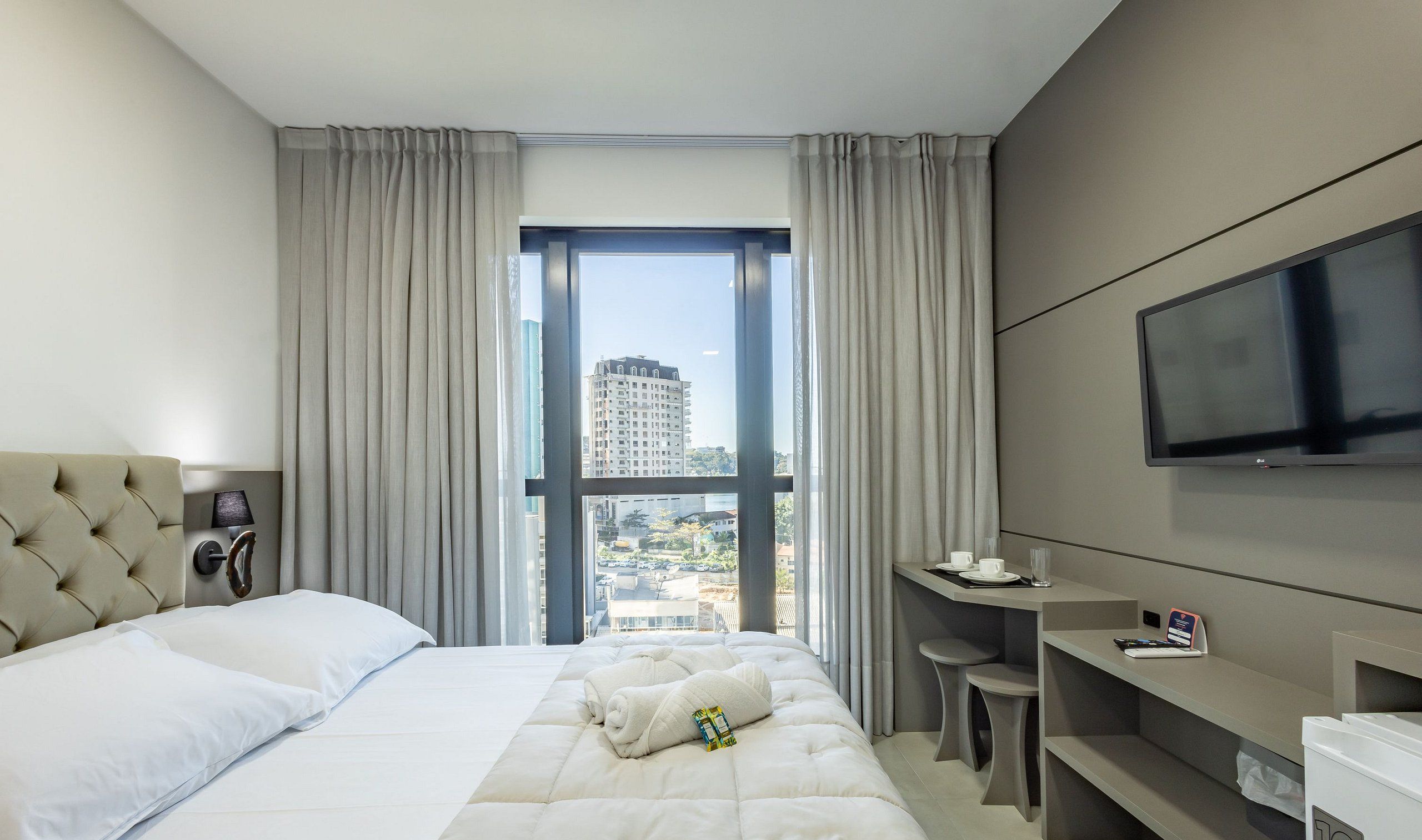 A hotel room with a large bed, a desk, a television, and a view of a cityscape through a large window.