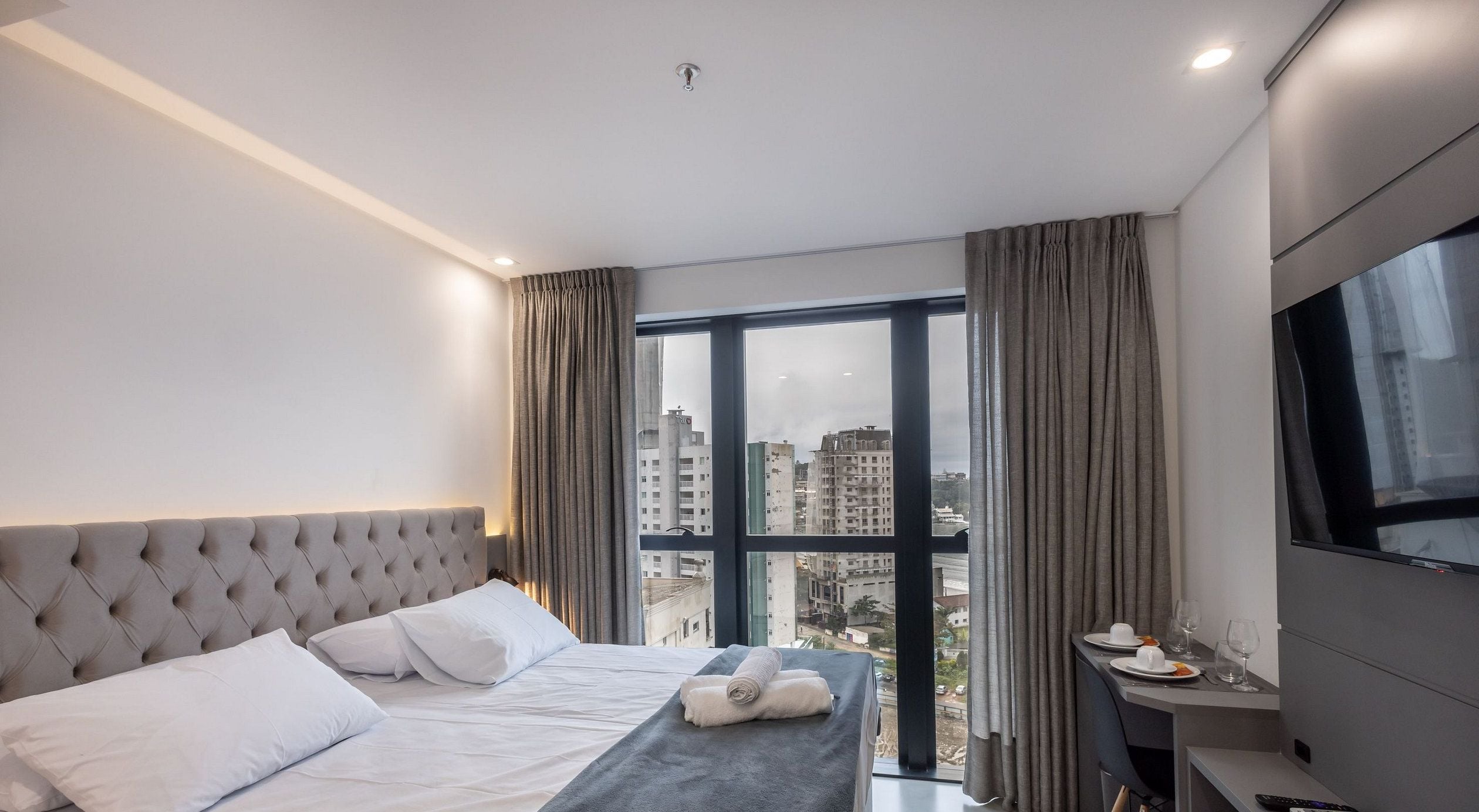A hotel room with a large bed, a desk with a TV, and a view of the cityscape through the window.
