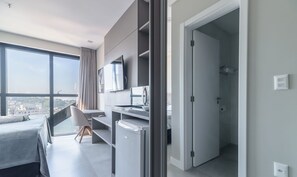 A modern hotel room with a bed, a desk, a television, and a view of the city. - MHFBs Edifício Moderno piscina vista mar (Itapema)