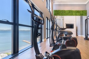Un gimnasio con equipo de ejercicio, ventanas amplias y vista al mar.
