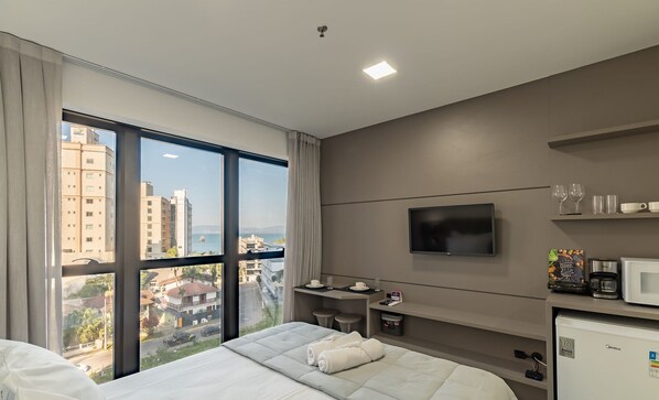 A hotel room with a large window, a bed, a TV, a minibar, and a view of the cityscape. - MHFBs Edifício Moderno piscina vista mar (Itapema)