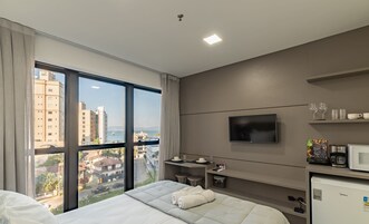 Habitación de hotel con un ventanal amplio, una cama, televisor, minibar y vistas a la ciudad.