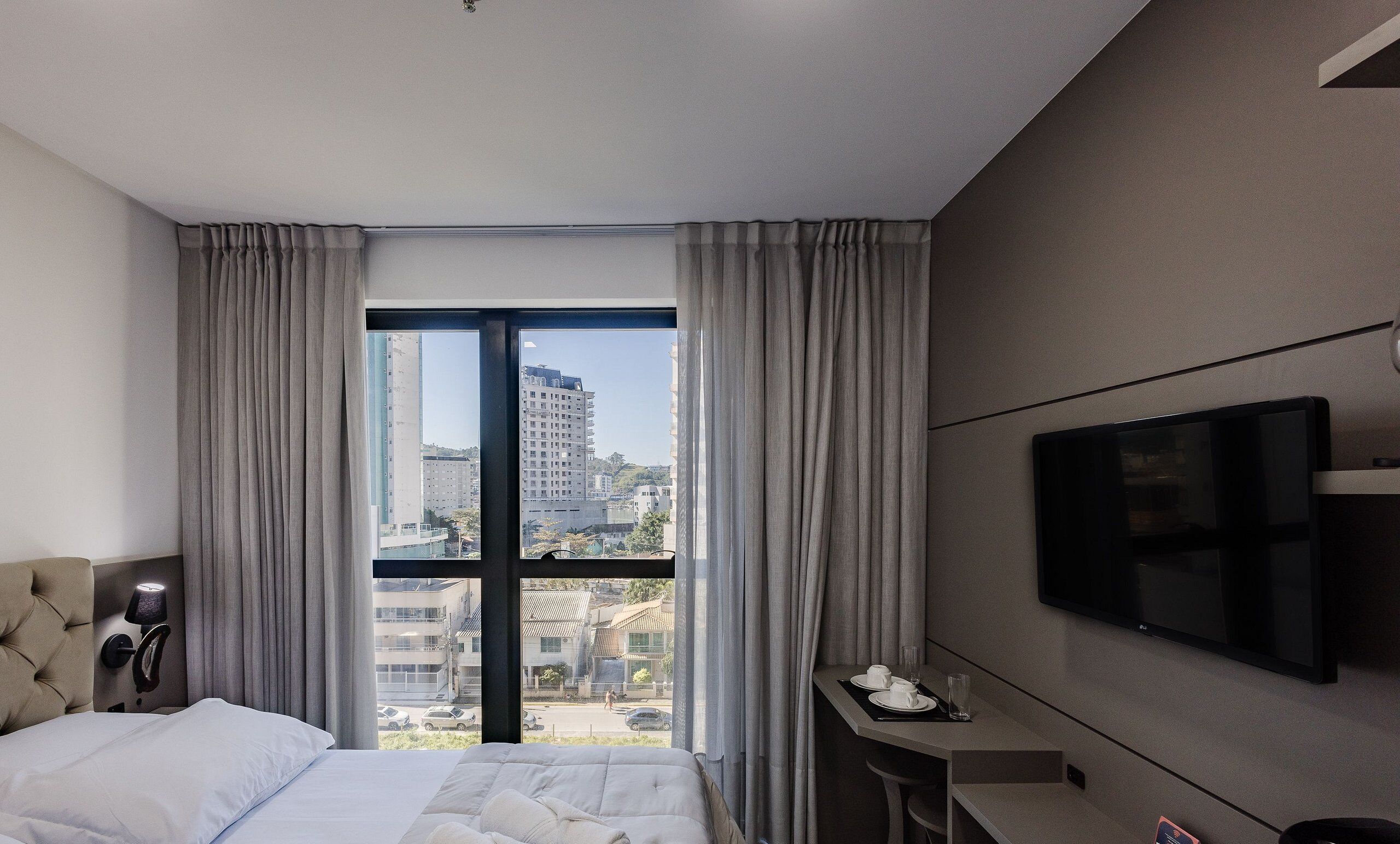 Une chambre d’hôtel moderne équipée d’un lit, d’une télévision, d’un bureau avec une tasse et un verre, et d’une fenêtre avec des rideaux.