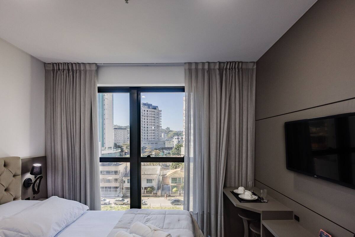 Une chambre d’hôtel moderne équipée d’un lit, d’une télévision, d’un bureau avec une tasse et un verre, et d’une fenêtre avec des rideaux.