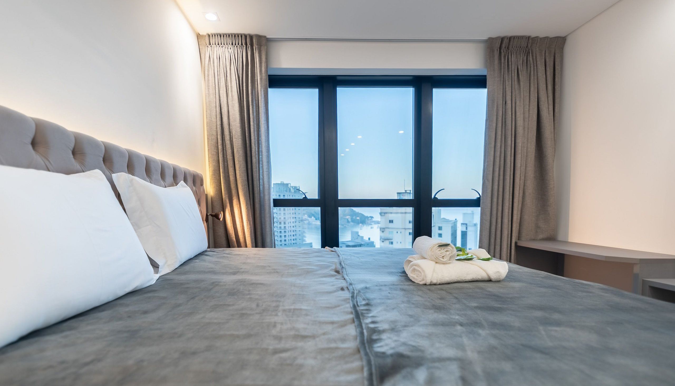 A hotel room with a large bed, a view of the city through floor-to-ceiling windows, and a small table with a towel.
