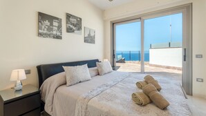 Un dormitorio con cama, almohadas, una mesita de noche, una lámpara y vistas al mar a través de una puerta corrediza de vidrio.