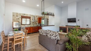 A modern kitchen with a built-in oven, a dining area with wooden chairs and a table, a sofa with patterned cushions, and a flat-screen TV mounted on the wall. - Holiday Home 'Casa Rural Adalid' with Pool, Garden, Terrace & Wi-Fi (La Puebla de Cazalla)