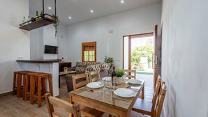 A dining area with a glass table, wooden chairs, a sofa, and a TV mounted on the wall. - Holiday Home 'Casa Rural Adalid' with Pool, Garden, Terrace & Wi-Fi (La Puebla de Cazalla)