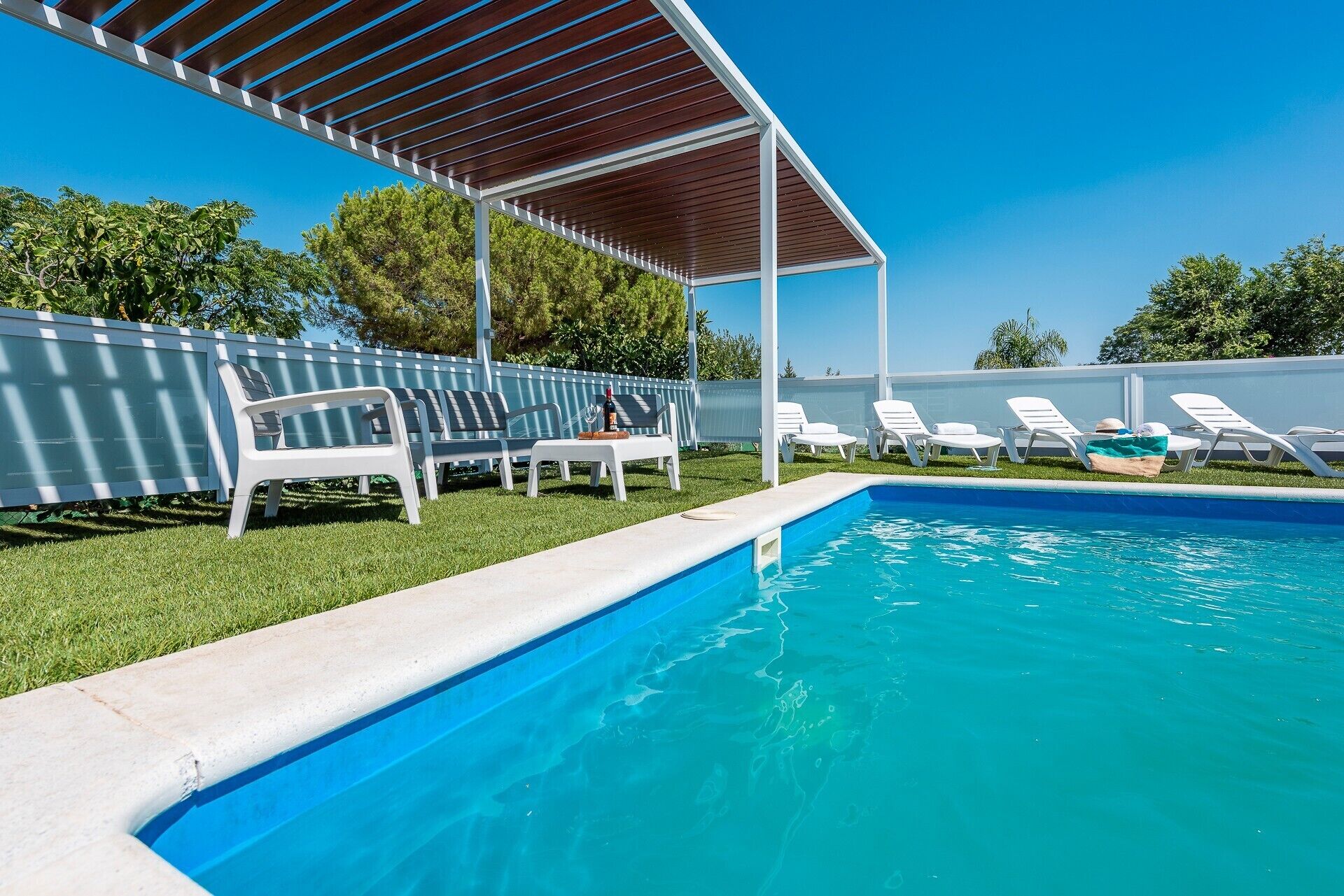 Piscina con una pérgola de madera sobre ella, rodeada de sillas reclinables blancas y un césped.