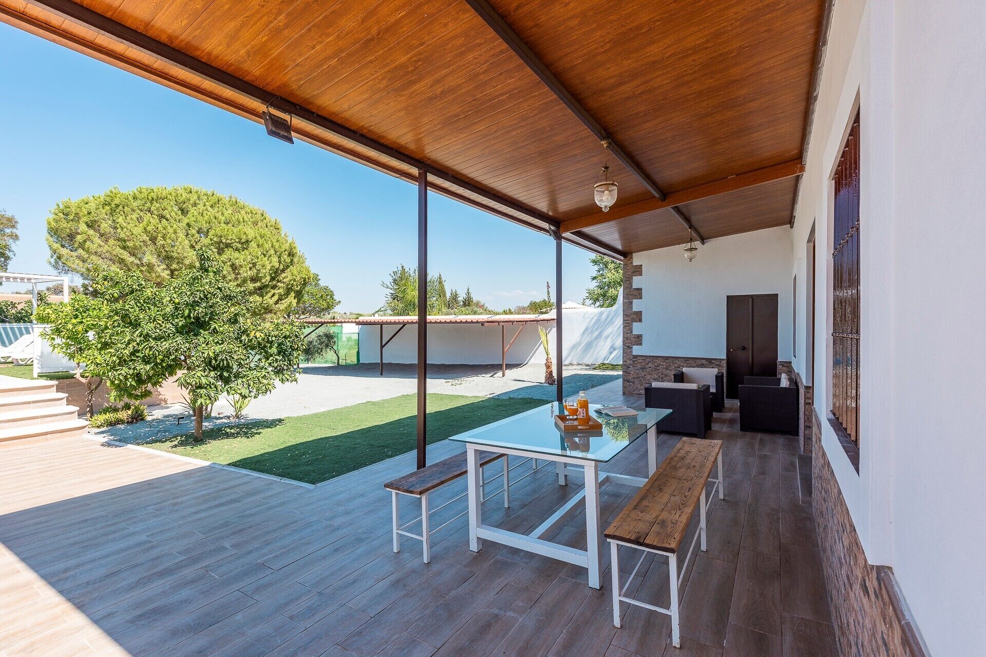 An outdoor patio with a glass table and benches, a wooden ceiling, and a view of a backyard with a tree and a white building.