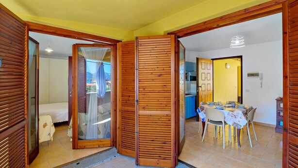 Una habitación con una cama, una puerta corredera de madera, una zona de comedor con mesa y sillas, y una cocina al fondo.