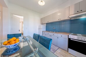 Une cuisine moderne avec des meubles blancs, un pare-feu en carreaux bleus, une table à manger en verre avec des chaises et un saladier rempli d’oranges.