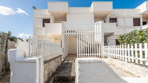 Un edificio blanco con entrada protegida por una reja, una escalera que conduce a un segundo piso y un cielo azul despejado.