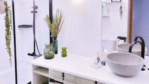 Un baño moderno con tocador blanco, un espejo grande, ducha con cabezal negro y una lámpara de diseño.