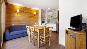 Ein gemütlicher Wohnraum mit Holzwand, einer Couch, einem Esstisch mit Stühlen, einer Küchenzeile und einem Fernseher auf einer Holzkommode.