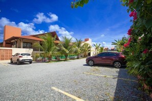 A parking area with two cars, a building with a red roof, and palm trees. - Viva Barra Hotel Pousada (Barra de São Miguel)
