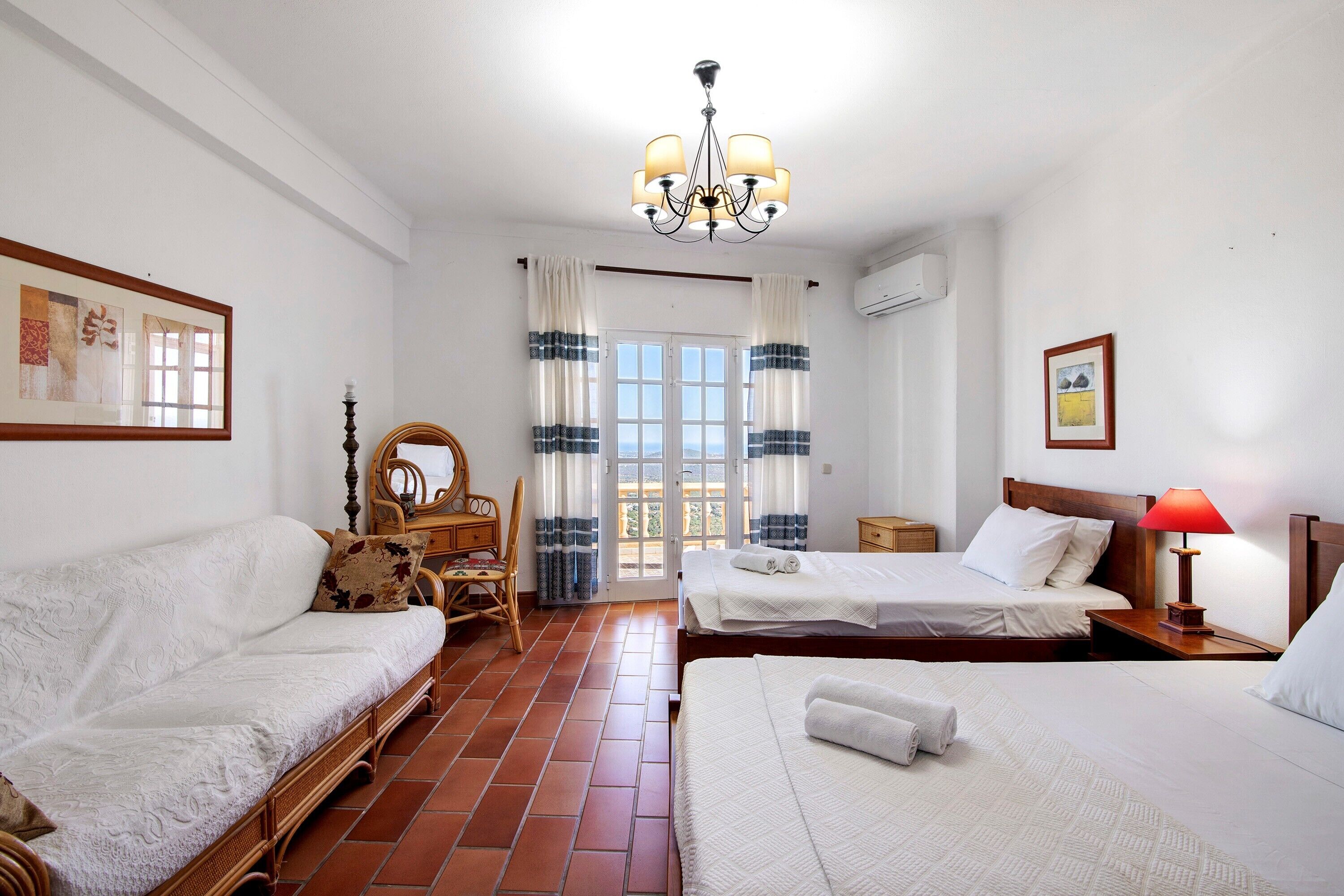 A hotel room with two beds, a sofa, a wooden chair, a chandelier, and a view through the window.