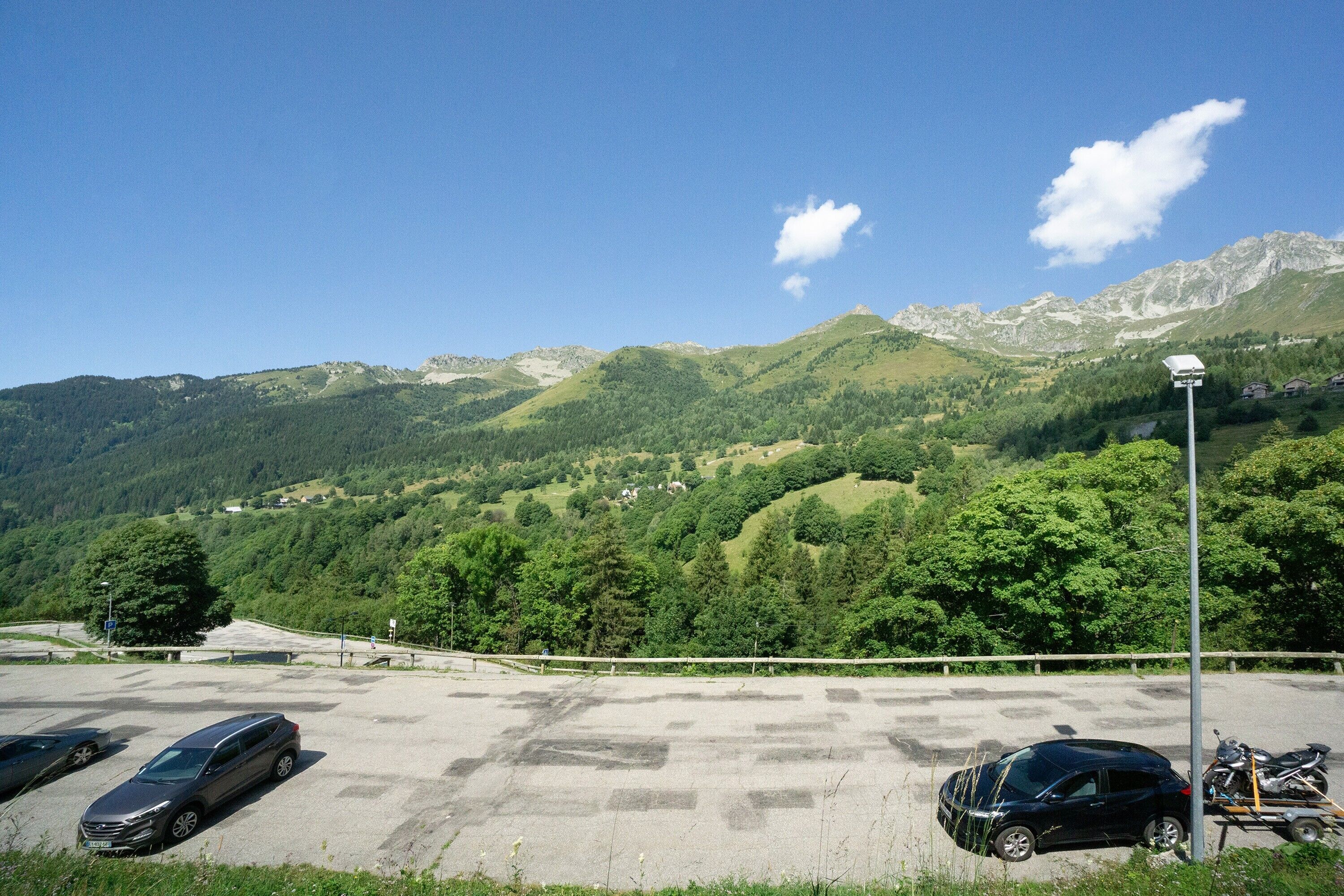 A parking lot with cars and a motorcycle, surrounded by green hills and mountains.