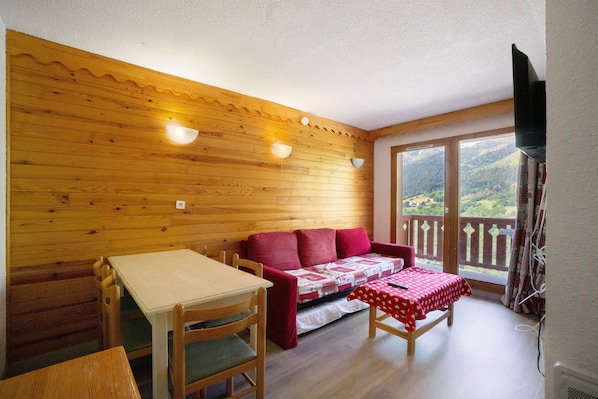 A room with wooden panel walls, a red sofa, a table with a red tablecloth, and a balcony with a view of the countryside.