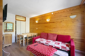 A cozy room with a wooden wall, a red sofa, a dining table with chairs, and a television mounted on the wall.