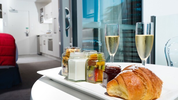 Un plateau de petit-déjeuner avec un croissant, des confitures de fruits et des coupes à champagne, posé sur une table dans une chambre d’hôtel moderne offrant une vue sur la ville.