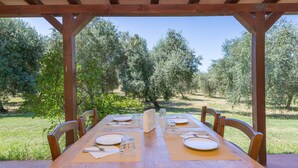 Une table en bois, dressée pour six personnes, avec des chaises, installée à l’extérieur et offrant une vue sur les oliviers.