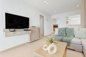 A modern living room with a flat-screen TV, a wooden coffee table with vases, a sofa with a throw blanket, and a kitchen area in the background.