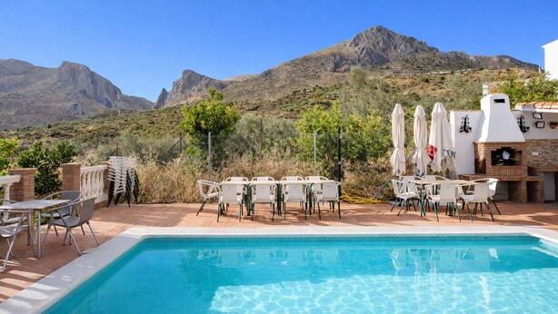 A poolside area with outdoor seating and a barbecue grill, surrounded by a mountainous landscape.