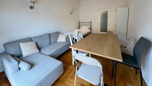 Une pièce avec un canapé gris, une table en bois, des chaises blanches, un lit et un tableau blanc.